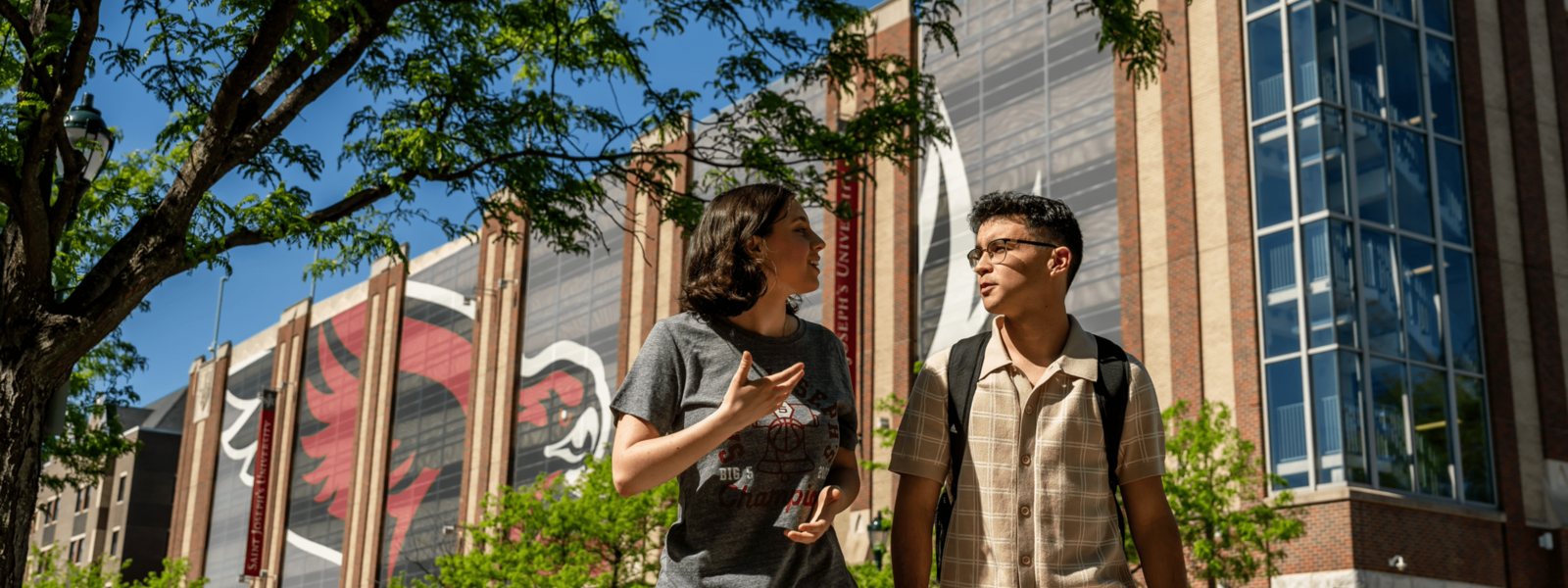 two students walk outside the hawk's landing parking garage with a large mesh hanging of the hawk logo behind them