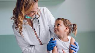 Nurse wearing a white lab coat and blue rubber gloves smiling at a young toddler sitting in a medical exam room