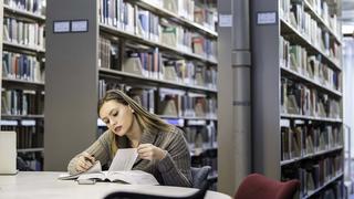 A woman sits at a table in the library looking at a book with stacks behind her