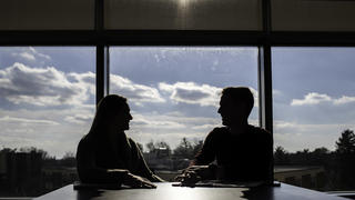 the shadows of two students can be seen talking in the library with bright windows behind them