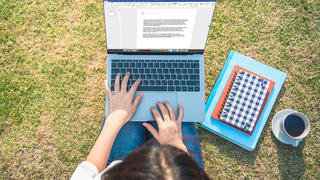 A person sits in the grass typing a document on their laptop. A notebook and coffee much is placed to the right of them.