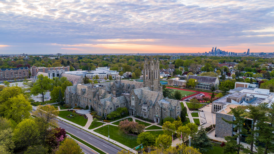 The green campus of Hawk Hill, Saint Joseph University's main campus, the gothic spires of Barbelin Hall rise in the foreground and the skyscrapers of Philadelphia loom in the background