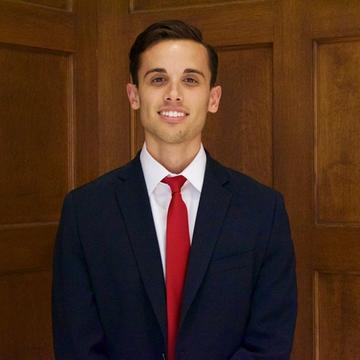 Nicholas smiles at the camera wearing a blue blazer, white shirt and red tie