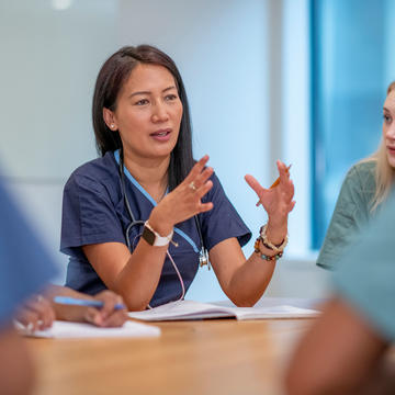 Healthcare professional wearing scrubs speaking to a group of nurses at a table