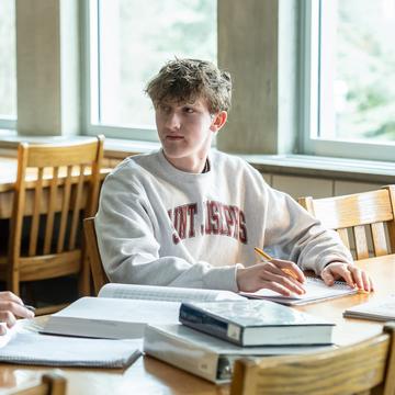 Student writing in a notebook and surrounded by books in a library