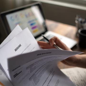 A hand holding a stack of resumes with a laptop in the background