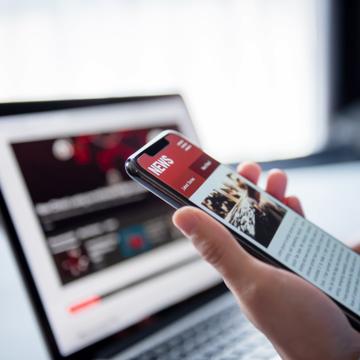 Person holding a phone with news articles on the screen with a laptop in the background