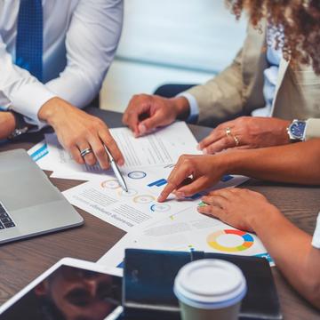 Zoomed in image of people pointing to papers with charts and graphs on a table with a tablet, computer, and cup of coffee