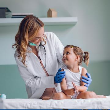 Nurse wearing a white lab coat and blue rubber gloves smiling at a young toddler sitting in a medical exam room