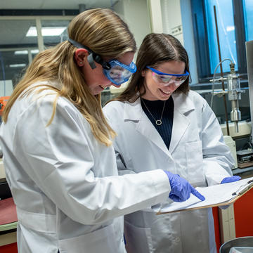 Two students wearing goggles in a lab pointing to a piece of paper