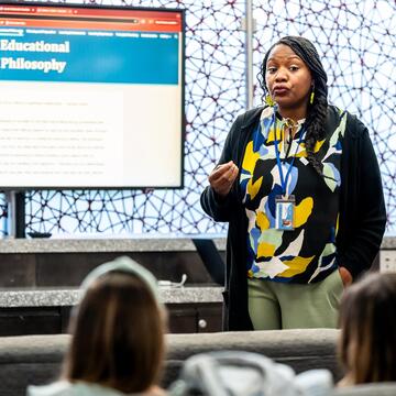 An educational leader delivering a presentation to a group of adults with a PowerPoint slide that says "Educational Philosophy"