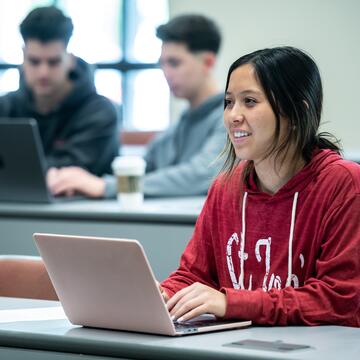 female student wearing red SJU sweatshirt typing on laptop in class 