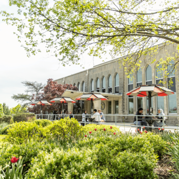 Wide shot of students sitting on picnic benches with umbrellas outside the Post Learning Commons