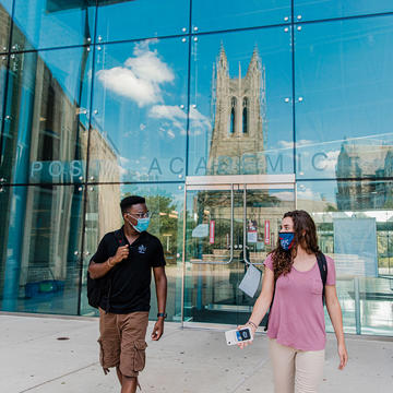 Two Saint Joseph's University students maintaining safe distance on campus wearing masks outside of the Drexel library with Barbelin Hall reflecting off the glass.