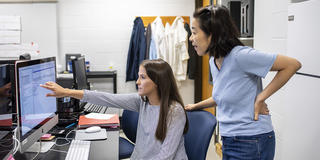 A faculty member leans behind a student who is working with data on a computer and pointing to it