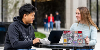 two students seated outdoors talking to each other over their laptops