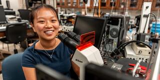 McNulty Scholar_Sarah Koh_Smiling at the camera from behind a microscope_2500x1400