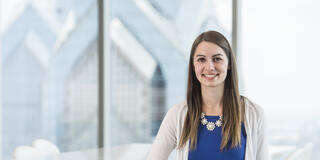 A student in a business casual outfit stands with a skyscraper behind her