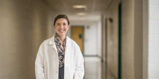 A woman in a white lab coat stands in a medical hallway smiling
