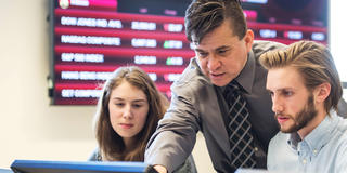 A professor stands behind two students looking at a computer with a trading room ticker behind them.