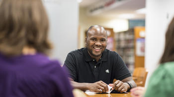 An adult student smiles at a table in the library wearing a polo