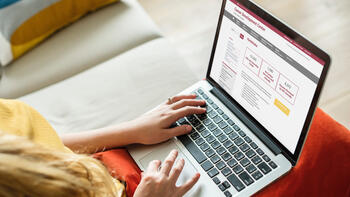 A woman sits on a couch with her laptop open. The laptop is displaying SJU's Career Development Center website.