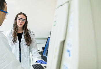 Student and faculty member wearing lab coats and safety goggles standing next to a computer
