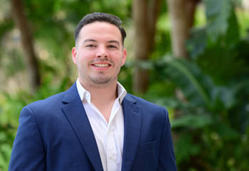 Headshot of Jake DeMarco in blue suit standing outside in front of trees
