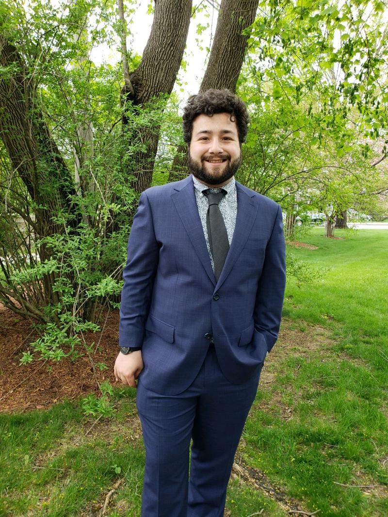 a young man outside, wearing a dark blue suit and smiling at the camera