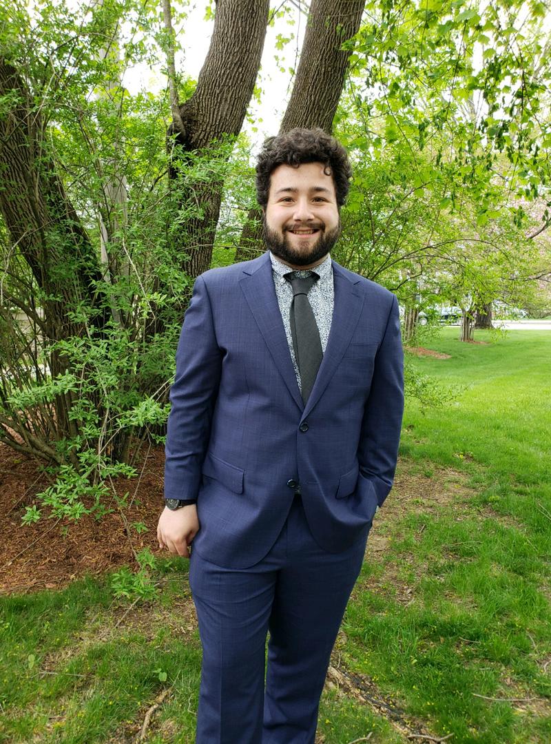 a young man outside, wearing a dark blue suit and smiling at the camera