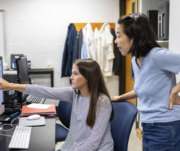 A faculty member leans behind a student who is working with data on a computer and pointing to it