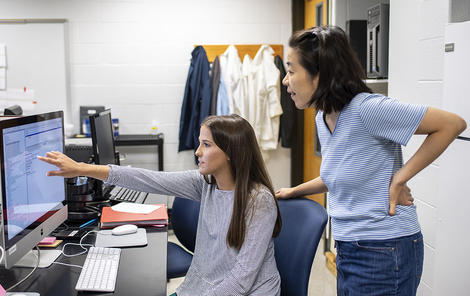 A faculty member leans behind a student who is working with data on a computer and pointing to it
