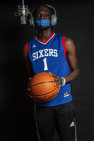 black man wearing a Sixers jersey with tape over his mouth at microphone