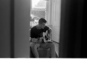 B+W photograph of man sitting on bed and playing guitar through doorway