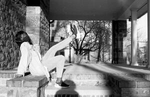 B+W photograph of woman in N95 mask sitting on steps and kicking leg out